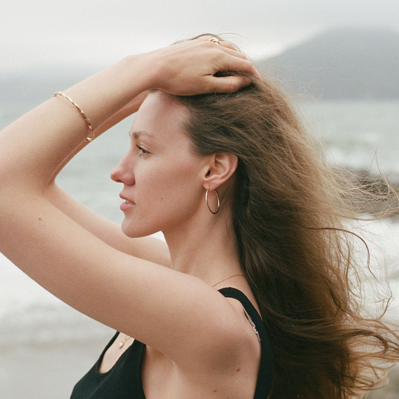A woman with long brown hair stands by the ocean, wearing a black top and gold hoop earrings, with her hand running through her hair against a blurred beach and water background.