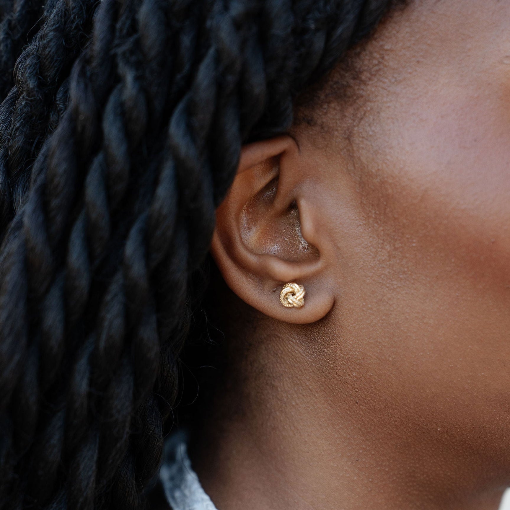A close-up side profile of a woman with black braided hair wearing a small gold stud earring.