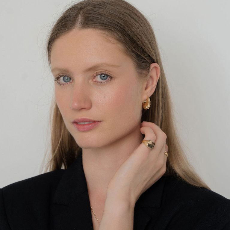 Woman wearing gold earrings and a ring, against a plain background