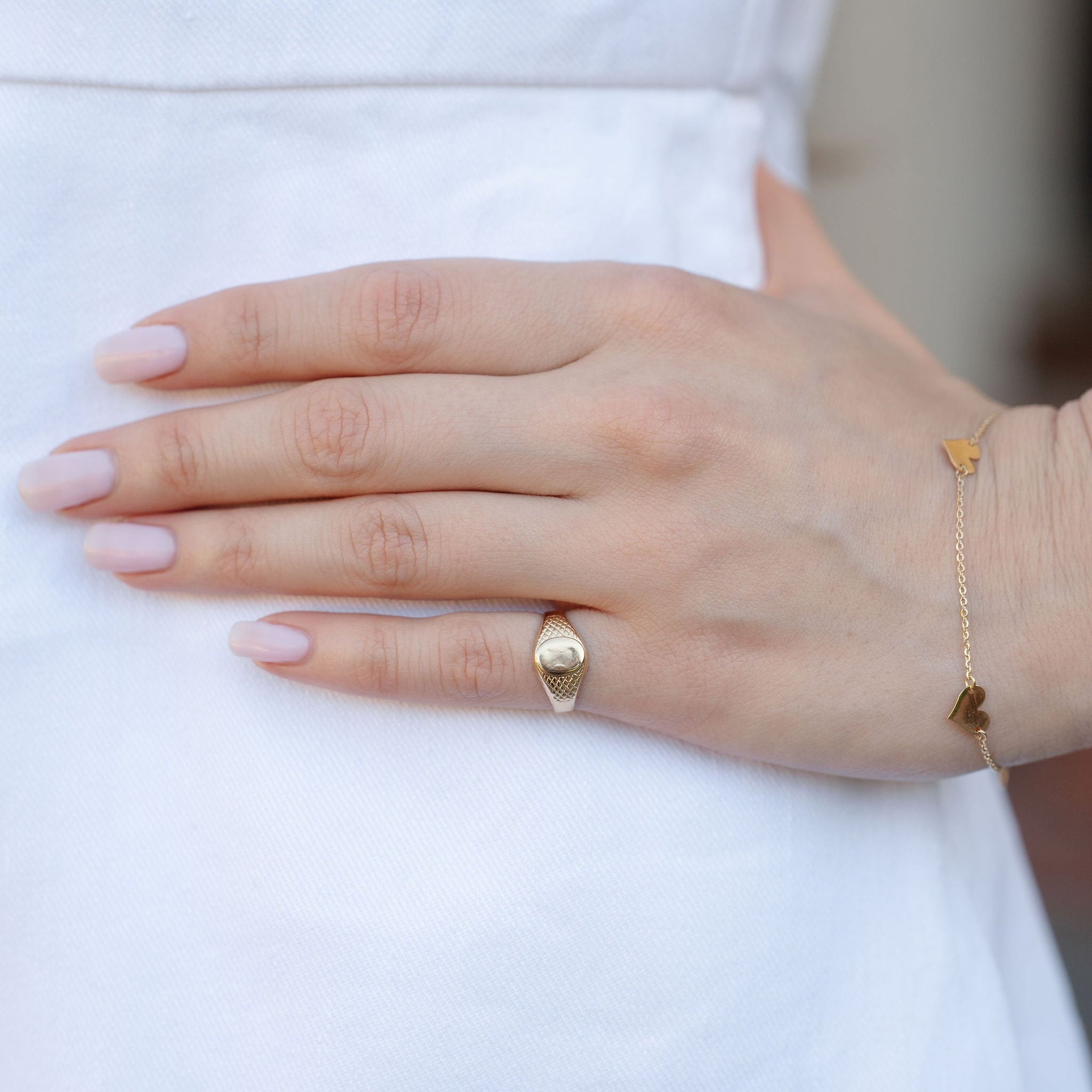 A close-up of a person's hand resting on a white dress, showcasing a gold signet ring with a small oval emblem on the ring finger and a delicate gold chain bracelet with heart charms on the wrist.