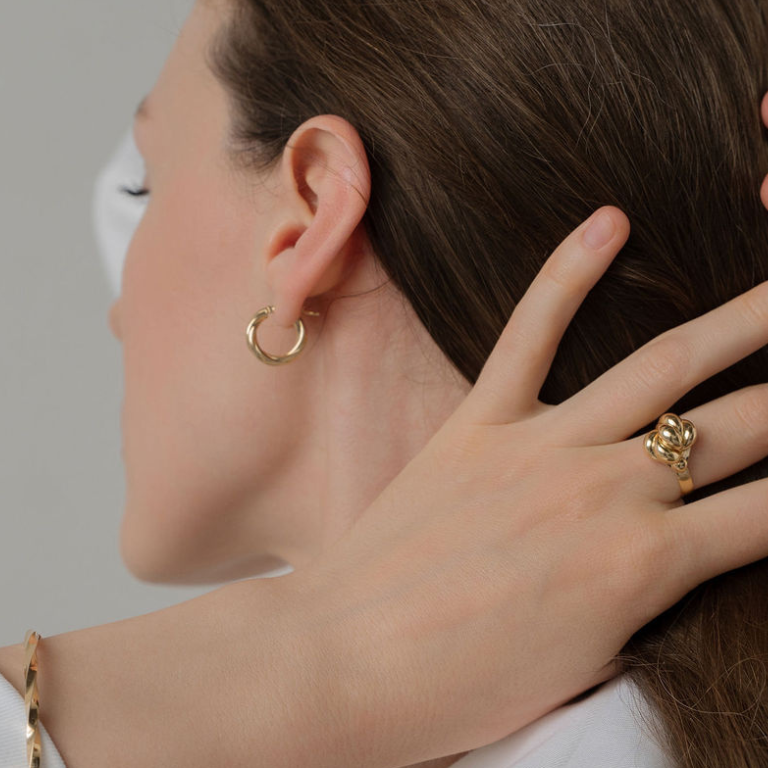 Close-up of a person wearing gold hoop earrings and a ring, with a neutral background.
