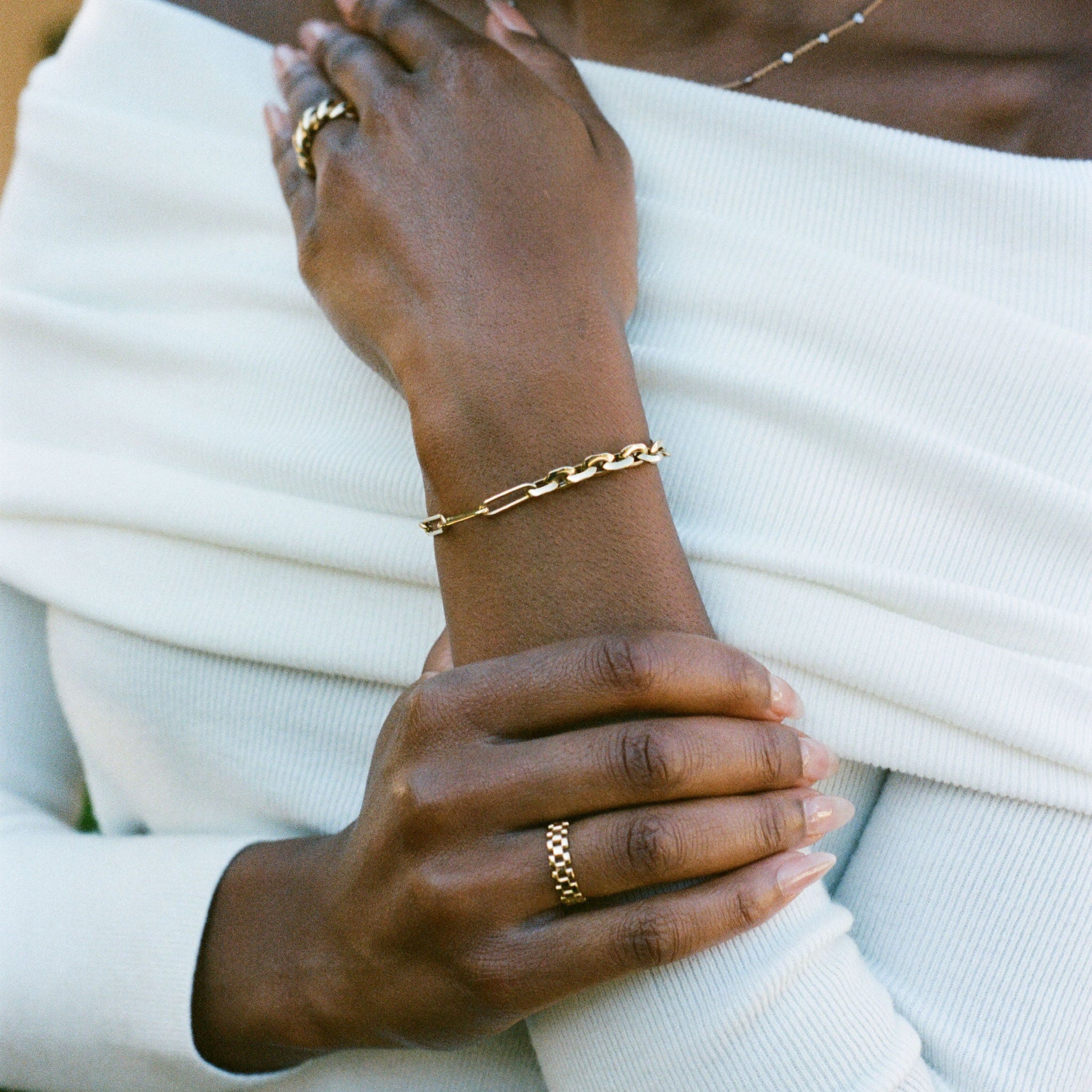 A close-up lifestyle shot of a model wearing a gold chain link bracelet and gold ring, styled with a white off-the-shoulder knit.