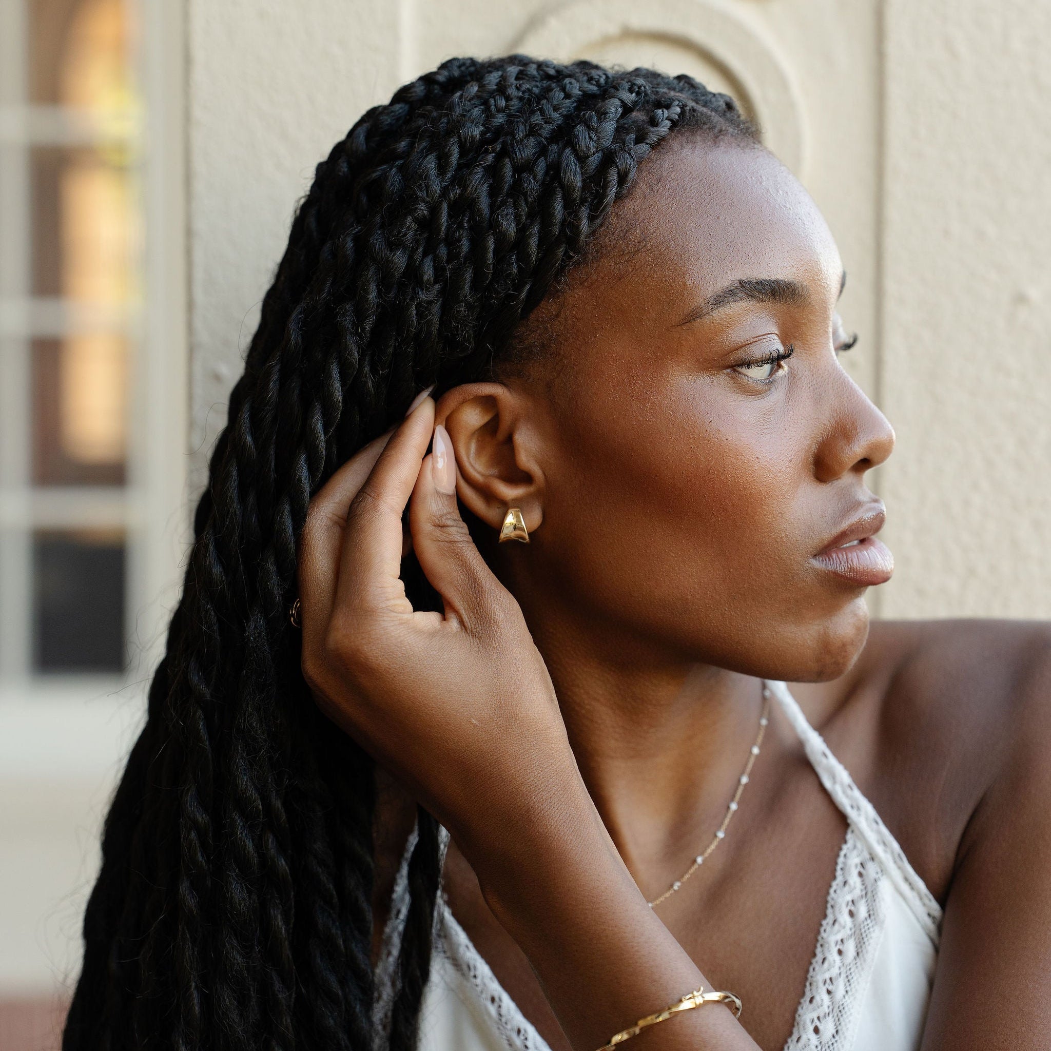 A woman with long black braided hair wearing a white lace-trimmed top and gold jewelry, posing in front of a beige stone wall.