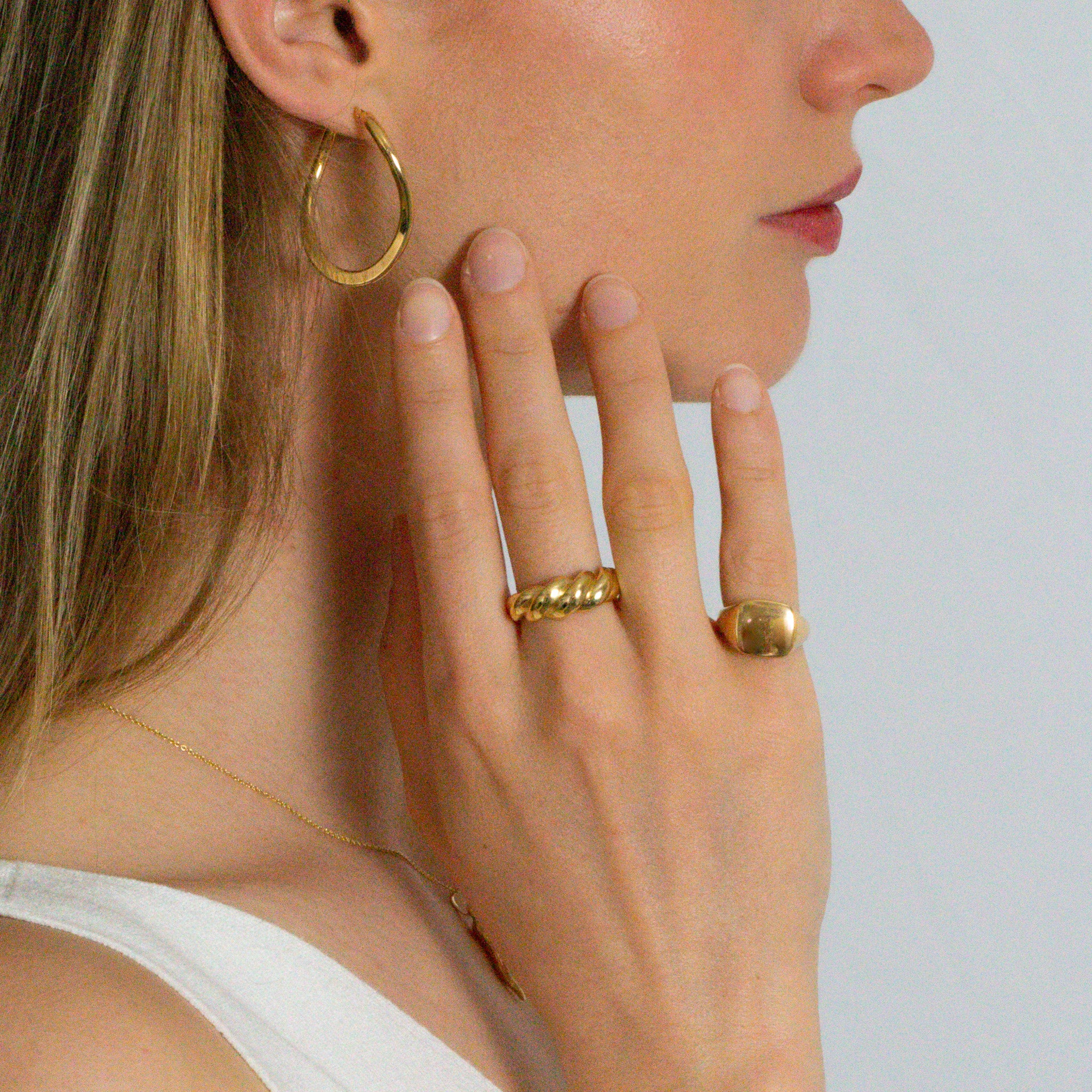 Close-up of a person wearing gold hoop earrings, rings, and a necklace on a plain background