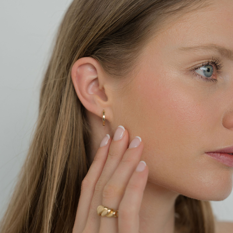 Close-up of a woman wearing gold hoop earrings and a ring, with a neutral background.