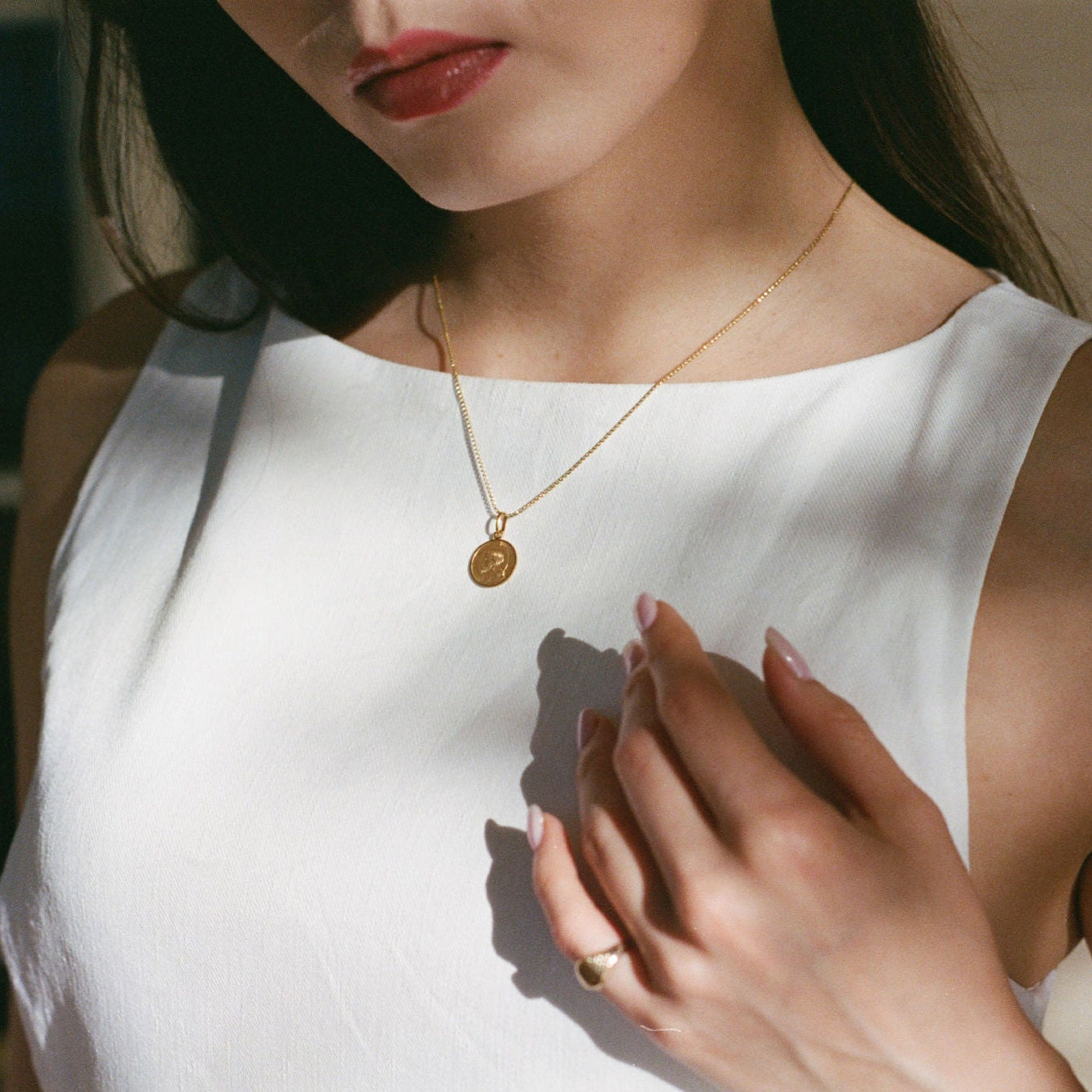 A close-up lifestyle shot of a woman wearing a dainty gold chain necklace with a round coin pendant under dramatic, high-contrast sunlight and shadows.