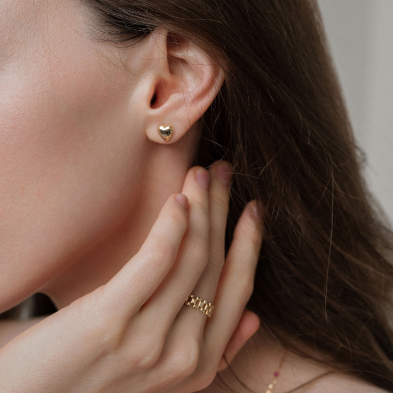 Close-up of a person wearing heart shape gold earrings and a ring, with a neutral background.