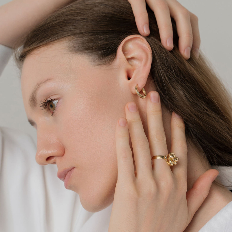 Close-up of a woman wearing gold hoop earrings and a ring, with a neutral background.
