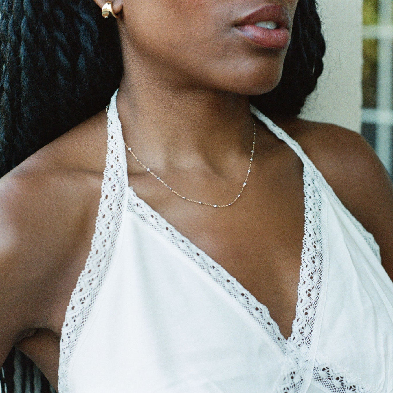 A close-up lifestyle shot of a woman wearing a white lace-trimmed halter top and a delicate gold necklace, with her dark braided hair and skin visible against a soft-focus background.